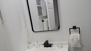 Un baño con un lavabo blanco, un grifo negro y un espejo encima. Se observa un perchero con una toalla blanca y un sujetatelas negro.