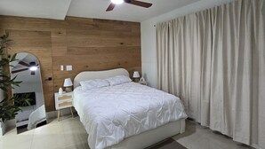 Un dormitorio con cama, mesitas de noche, ventilador de techo, espejo y ventana con cortinas.