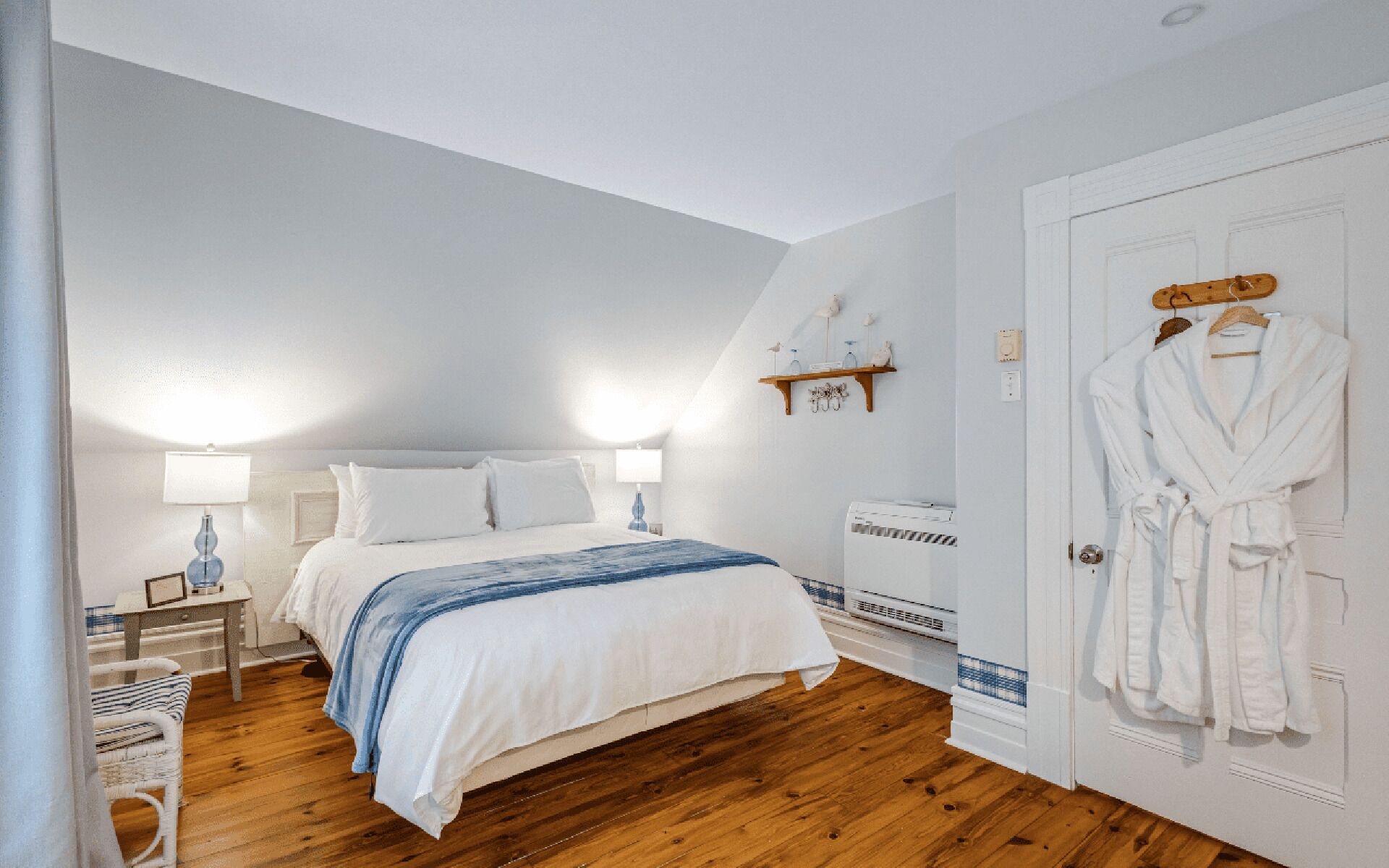 Une chambre à coucher avec un lit, des tables de chevet, une lampe, un radiateur, un peignoir accroché à la porte et une étagère avec des objets décoratifs.