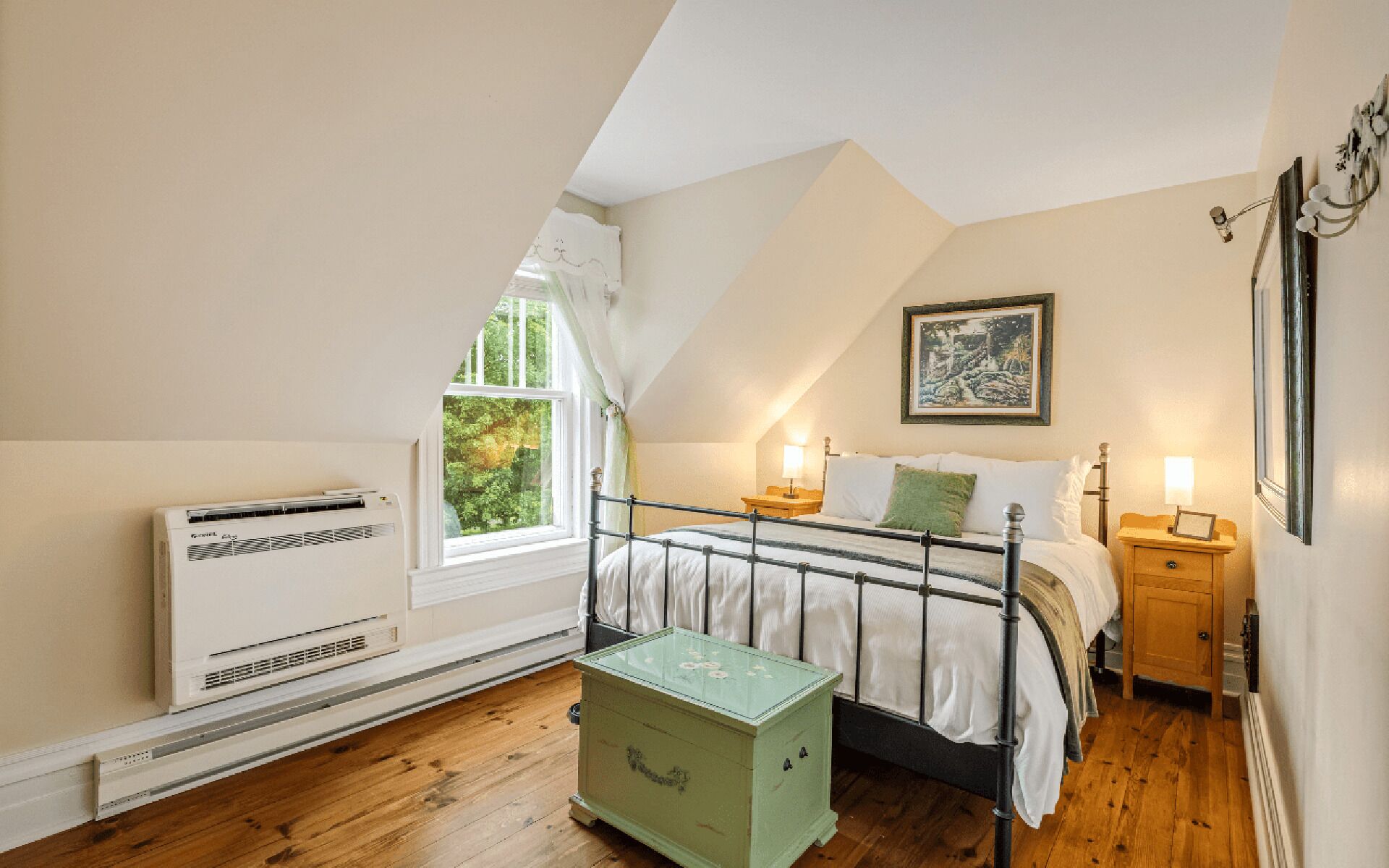 Une chambre avec un plafond en pente, un lit avec une literie blanche, une commode verte, un radiateur et une fenêtre donnant sur des arbres.