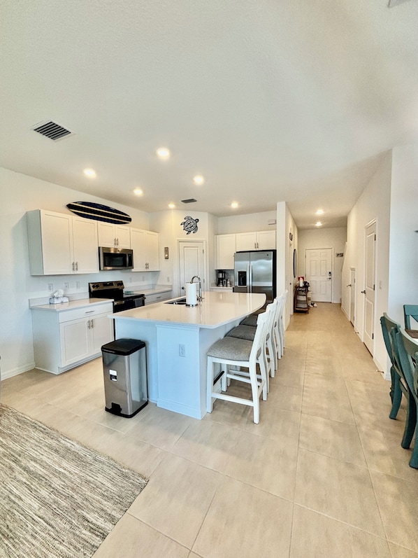 Una cocina moderna con muebles blancos, una isla central, electrodomésticos de acero inoxidable y suelo de baldosas.