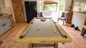 A pool table with a billiard cue, a black refrigerator, and a wooden cabinet in a room with a tiled floor and a view of the outdoors. - Wood Farm Cottage (Boston)