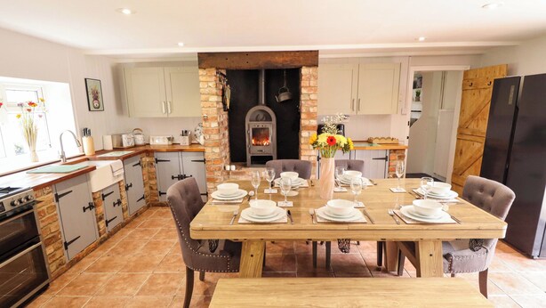 A modern kitchen with a central island, a dining table set for six, and a wood-burning stove. - Wood Farm Cottage (Boston)