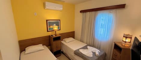A hotel room with two single beds, a nightstand with a lamp, an air conditioner, a framed picture, and a window with curtains.