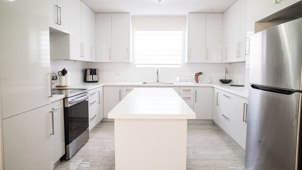 Une cuisine moderne avec des meubles blancs, un îlot central, des appareils électroménagers en acier inoxydable et une fenêtre qui laisse entrer la lumière naturelle.