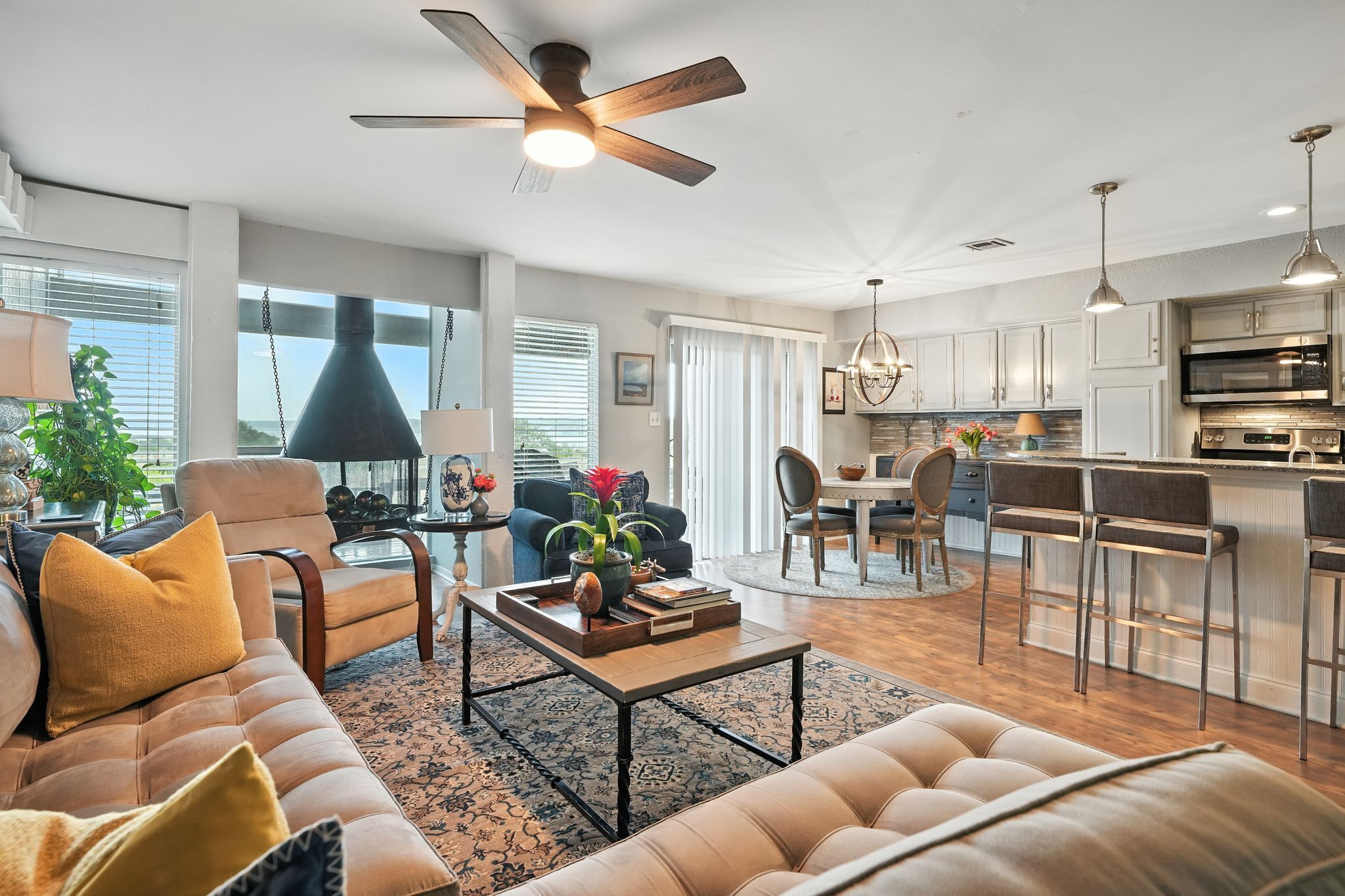 Una sala moderna con sofá, sillón y una mesa de centro, un comedor con candelabro, y una cocina con taburetes de bar y electrodomésticos de acero inoxidable.