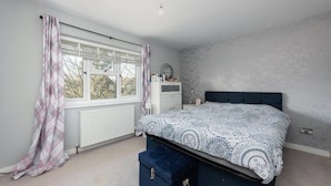 A bedroom with a bed, a window with curtains, a radiator, a bedside table, and a wall-mounted clock. - The Willow - Home with modern facilities in the outstandingly beautiful Surrey (Woking)