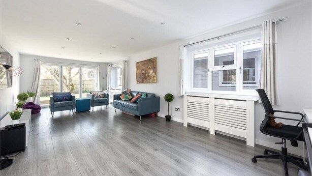 A spacious living room with a grey wooden floor, a blue sofa, a black office chair, and a white radiator. - The Willow - Home with modern facilities in the outstandingly beautiful Surrey (Woking)