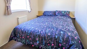 A bedroom with a bed, a window with curtains, a bedside table, and a radiator. - Willerby Aurora B14 (Abergele)