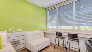 Une pièce moderne avec un mur vert, un canapé blanc, deux tabourets de bar et une fenêtre avec des stores.