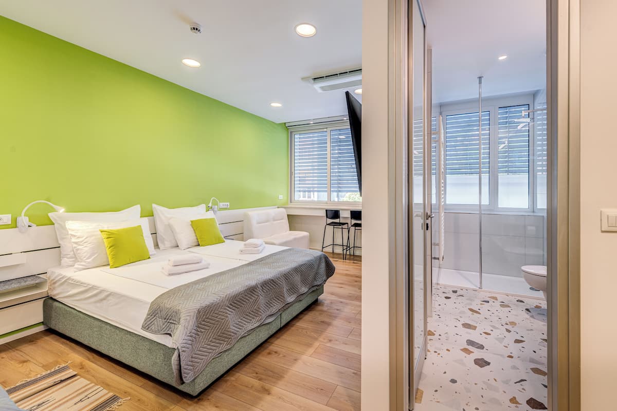 Un dormitorio moderno con una pared de acento verde, una cama grande con ropa de cama blanca y verde, una zona de ducha con puerta de cristal y un pequeño rincón de descanso con escritorio y sillas.