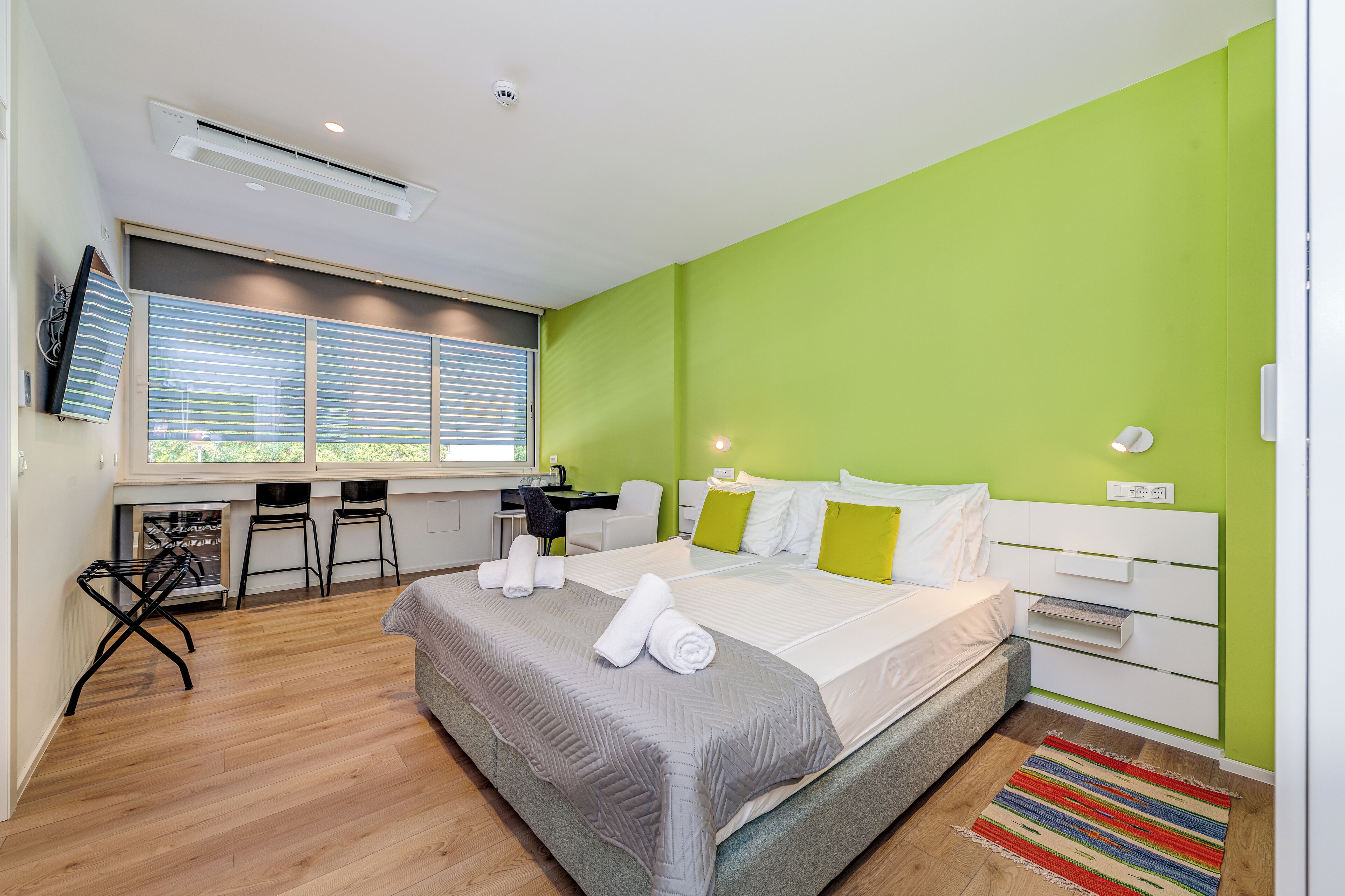 A modern hotel room with a large bed, a desk, and a window with blinds.