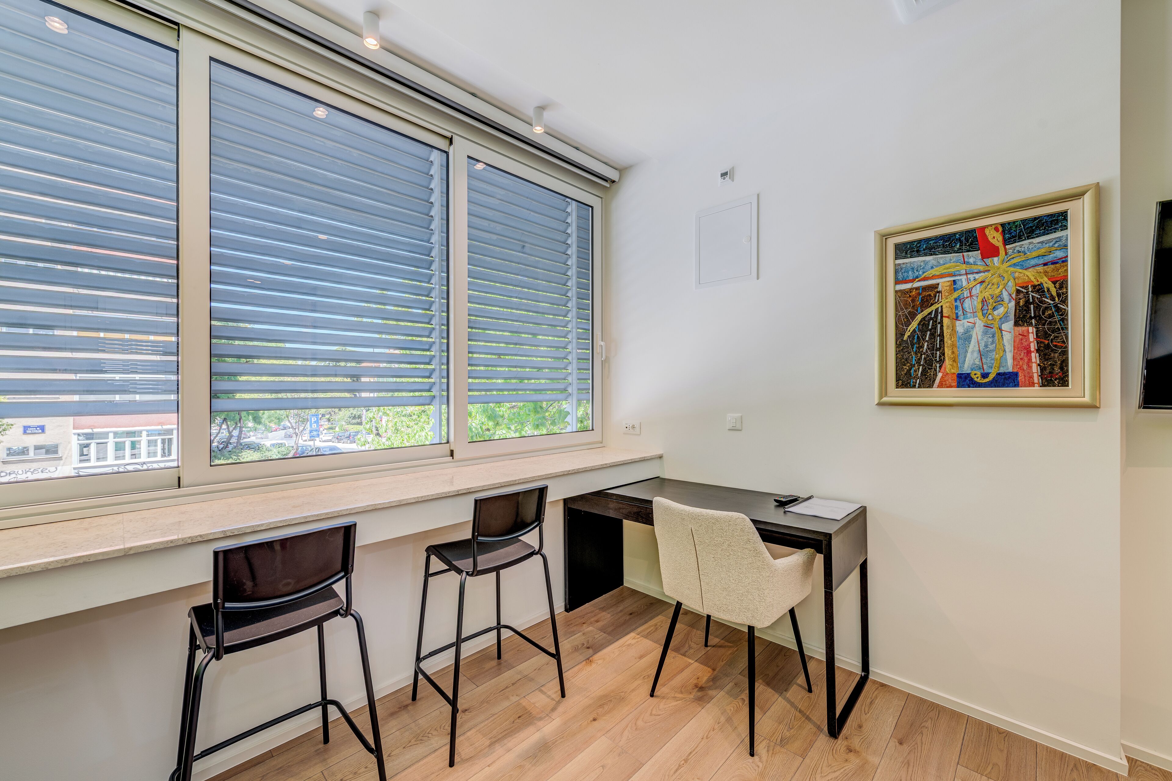 A modern room with a desk, two chairs, a large window with blinds, and a framed artwork on the wall.