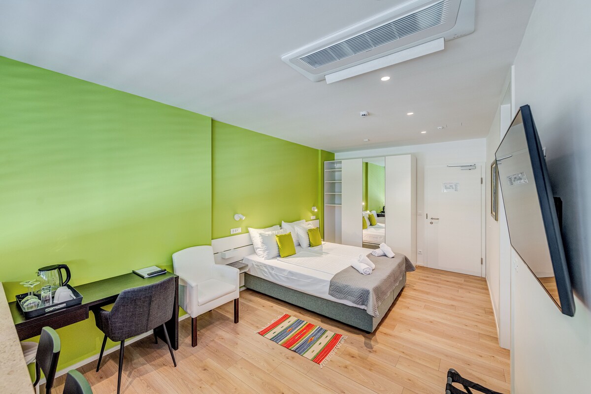 A modern hotel room with a green accent wall, a bed with white linens, a desk with a chair, a TV mounted on the wall, and a wooden floor.