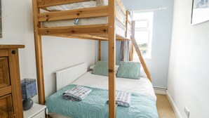 Una cama litera con estructura de madera, un colchón blanco y una colcha color aguamarina. Debajo de la litera inferior, hay un radiador blanco, una mesita de noche de madera con una lámpara y una ventana con cortinas.