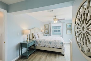 A bedroom with a bed, nightstand, ceiling fan, and wall art.