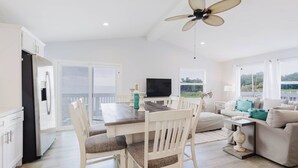 A modern kitchen with white cabinetry, a dining table with chairs, a ceiling fan, and a TV.