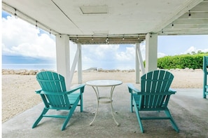 Dos sillas Adirondack color turquesa y una mesa redonda pequeña bajo un porche cubierto, con vista a la playa y al océano.