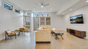 Un salon spacieux avec un canapé beige, deux fauteuils en cuir, une table basse et un meuble en bois. De grandes fenêtres sont équipées de stores, et on y trouve également un ventilateur de plafond et un tableau accroché au mur.