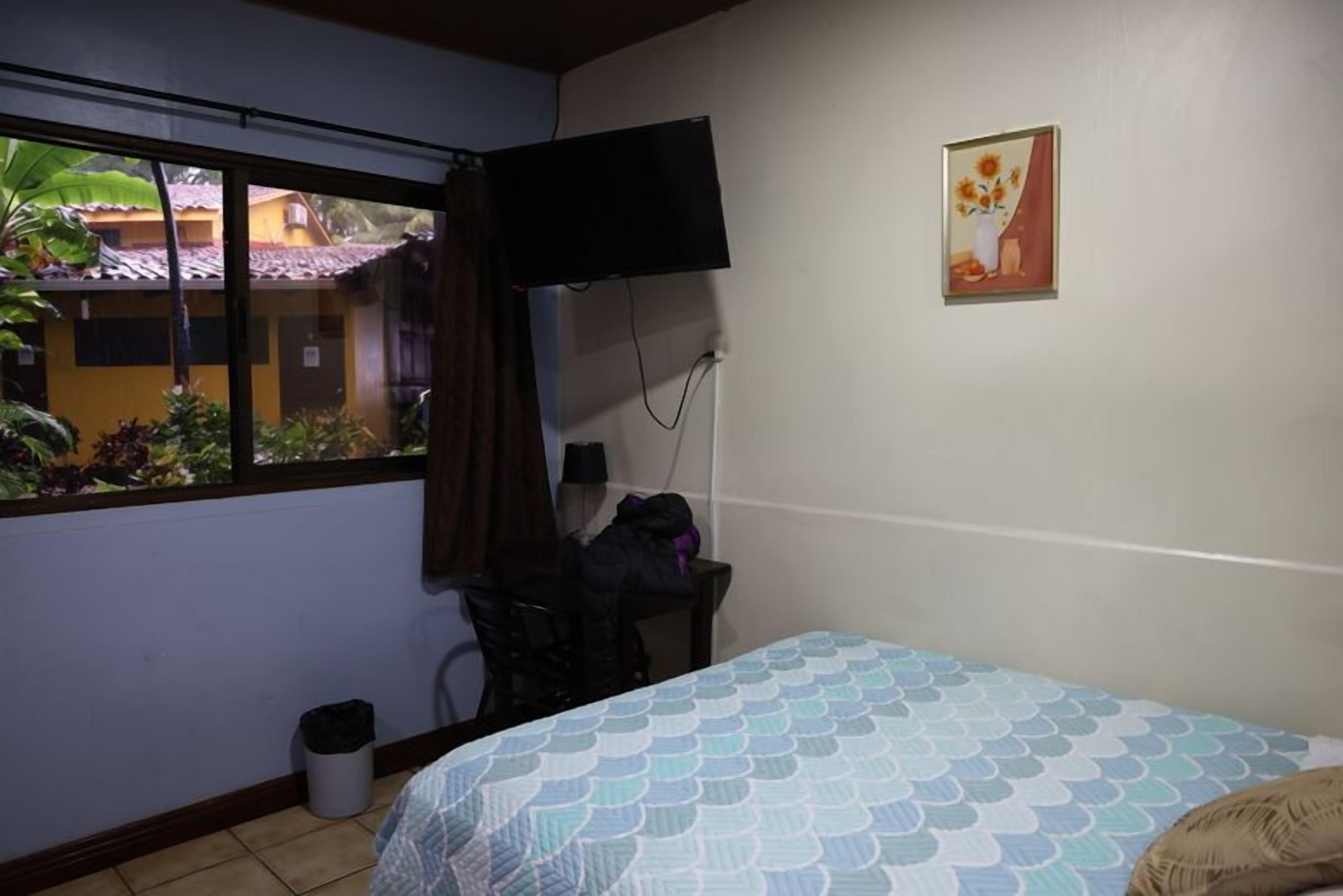 Une chambre avec un lit, un téléviseur fixé au mur, une fenêtre donnant sur les bâtiments environnants et une petite poubelle.