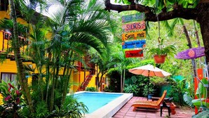 A pool area with a wooden bench and umbrella, surrounded by lush greenery and a yellow building. - Hotel Brasilito (Cabo Velas)