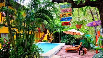 A pool area with a wooden bench and umbrella, surrounded by lush greenery and a yellow building.