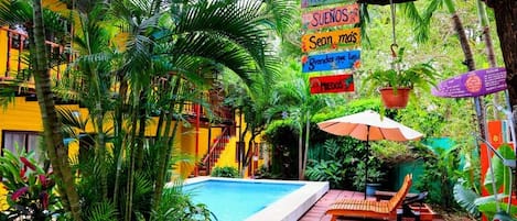 A pool area with a wooden bench and umbrella, surrounded by lush greenery and a yellow building.