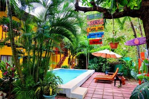 Un área de piscina con un banco de madera y una sombrilla, rodeada de abundante vegetación y un edificio amarillo.