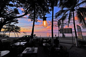 Un espacio al aire libre con mesas y sillas, con vista a una playa y un atardecer de fondo.