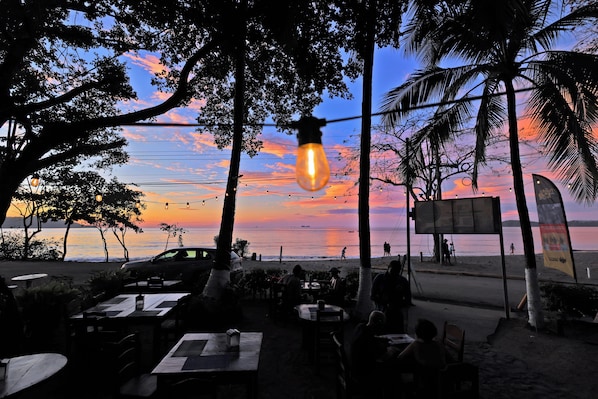 Uma área de estar externa com mesas e cadeiras, com vista para uma praia e um pôr do sol ao fundo.