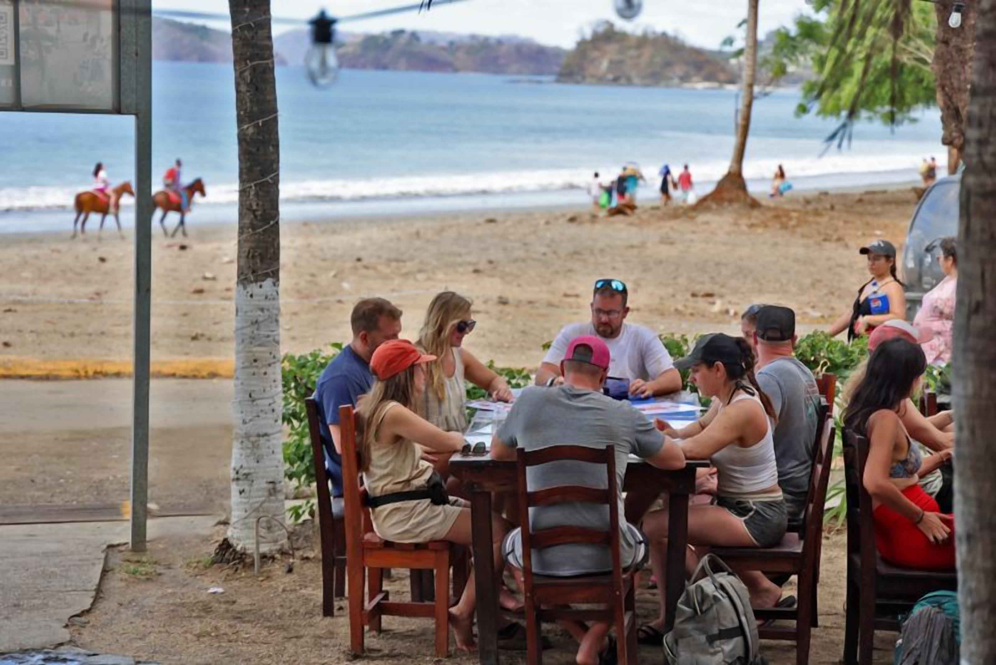 A group of people seated at a wooden table outdoors, with a beach and ocean in the background.