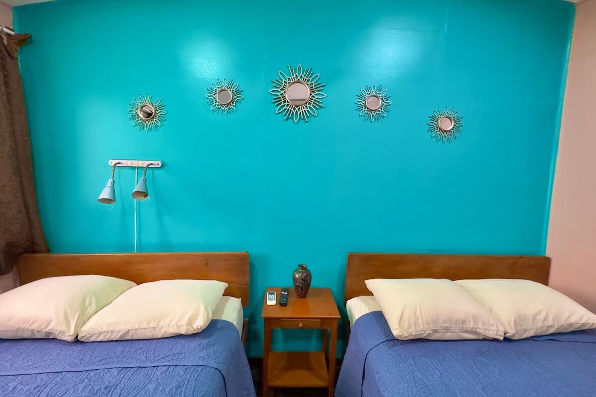 A room with two beds, a nightstand, a wall-mounted lamp, and decorative sun-like wall art.