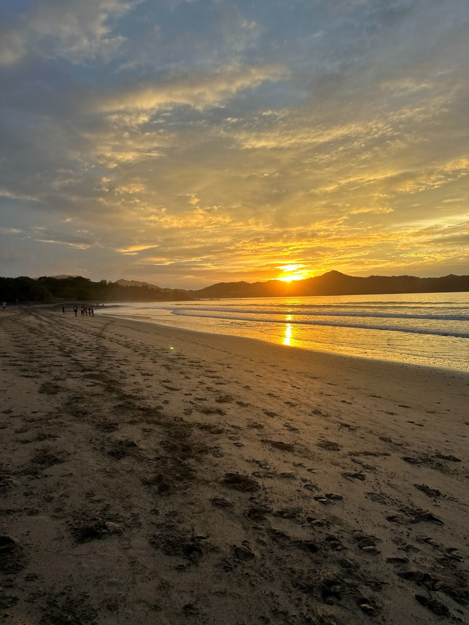 Uma praia com pegadas que levam à água, um pôr do sol ao fundo e uma cadeia montanhosa distante.