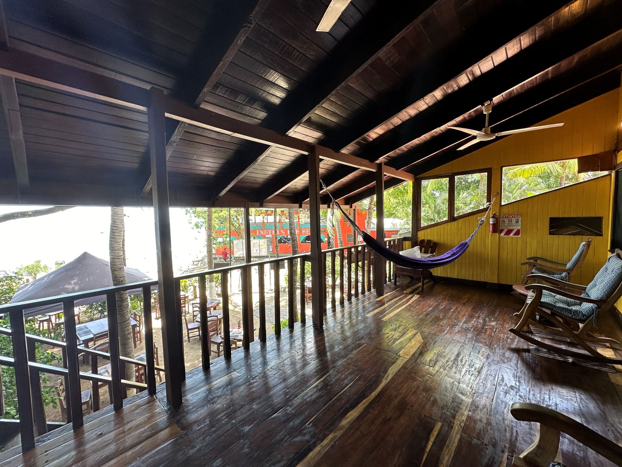 A wooden deck with a hammock and rocking chairs, overlooking a street with parked cars and buildings.