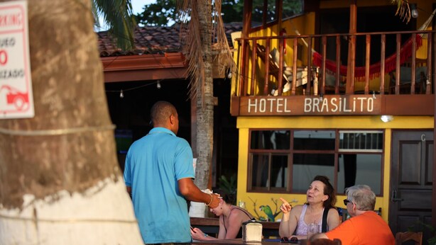 A hotel named 'HOTEL BRASILITO' with a signboard, a palm tree, and a wooden table with people seated. - Hotel Brasilito (Cabo Velas)
