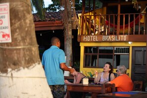 Un hotel chiamato 'HOTEL BRASILITO' con una insegna, una palma e un tavolo di legno con persone sedute.