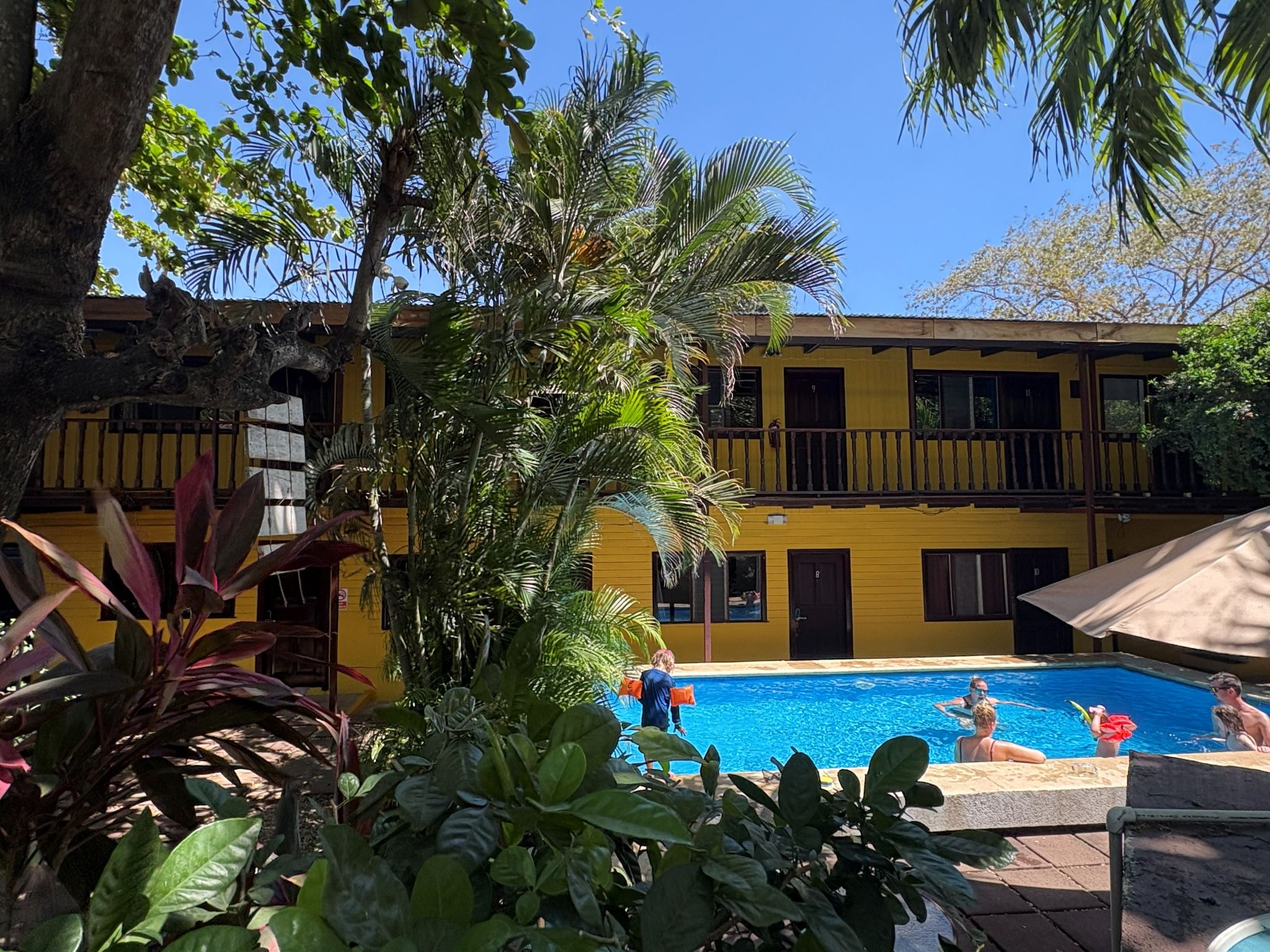 A pool area with a yellow building, palm trees, and a clear blue sky.