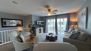 A living room with a TV, seating, a ceiling fan, and a view to the outside.