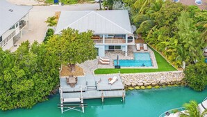 A two-story house with a pool, dock, and lush greenery.