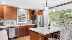 A modern kitchen with wooden cabinets, a stainless steel refrigerator, a built-in oven, and a large window with blinds.