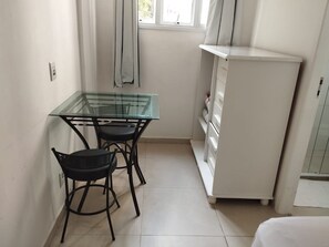 Ein kleines Zimmer mit einem Glastisch, zwei Stühlen, einem weißen Schrank und einem Fenster mit Vorhängen.