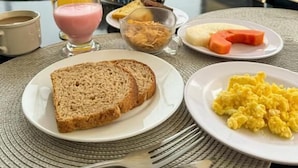 Desayuno con tostadas, huevos revueltos, un vaso de jugo de naranja y una taza de café sobre una mesa con vista a edificios.