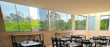 A dining area with multiple tables and chairs, large windows offering a view of greenery and buildings.