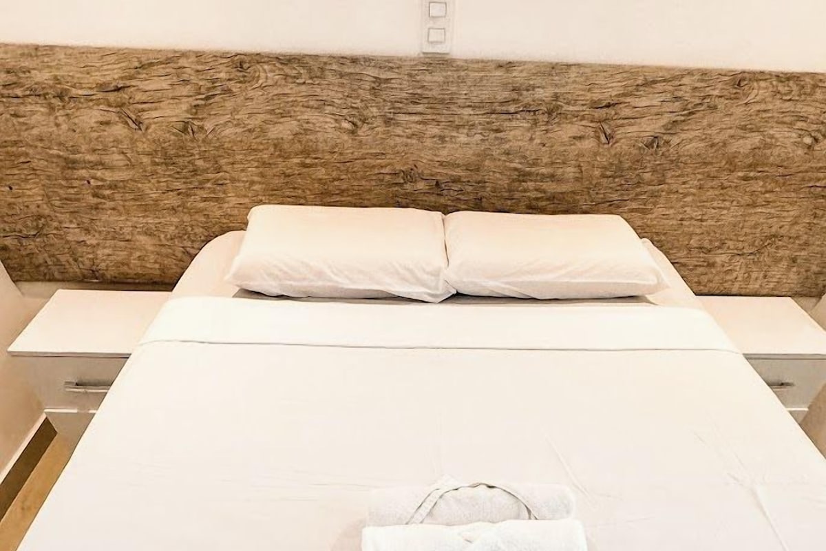 Ein ordentlich bezogenes Bett mit gefaltetem Handtuch, einem hölzernen Kopfteil und einer weißen Wand mit Lichtschalter.