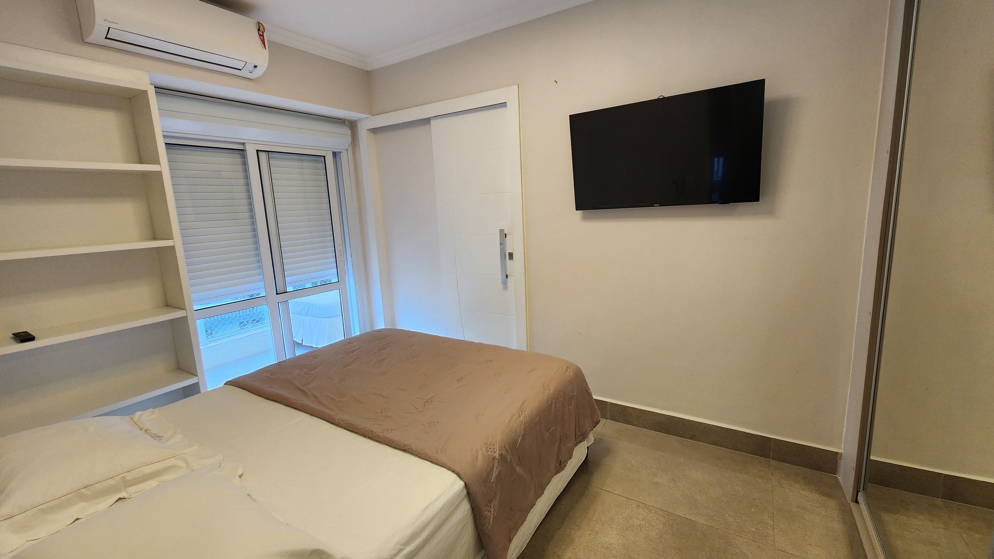 Un dormitorio con cama, televisor montado en la pared, armario y ventana con persianas.
