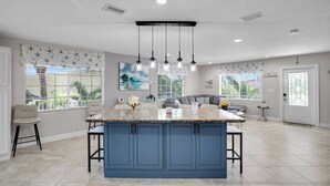 Una cocina amplia con una isla azul, iluminación colgante y ventanas grandes con persianas.