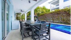 Un área de comedor al aire libre techada con piscina, ventiladores de techo y vista a un edificio blanco.