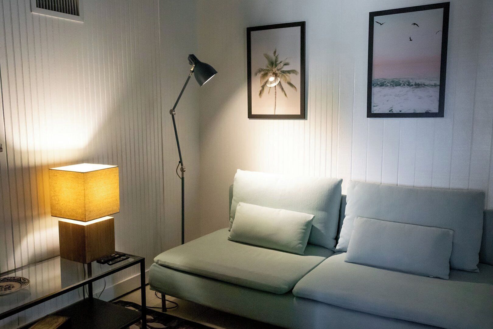 Una habitació amb un llit, una làmpada de peu, un quadre amb una palmera i un altre quadre amb una escena de platja.