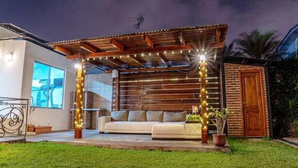 Un espacio de estar al aire libre cubierto, con un pérgola de madera, un sofá y luces colgantes.