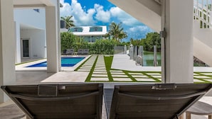 Uma área de piscina com espreguiçadeiras, céu claro e um edifício ao fundo.